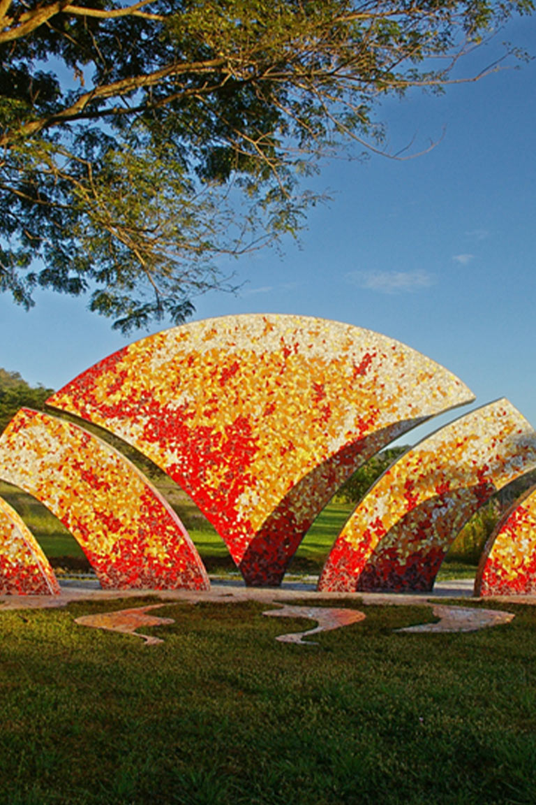 Escultura transitable de grandes dimensiones compuesta de fragmentos de mosaico de color rojo, naranja, amarillo y blanco, en medio de un parque.