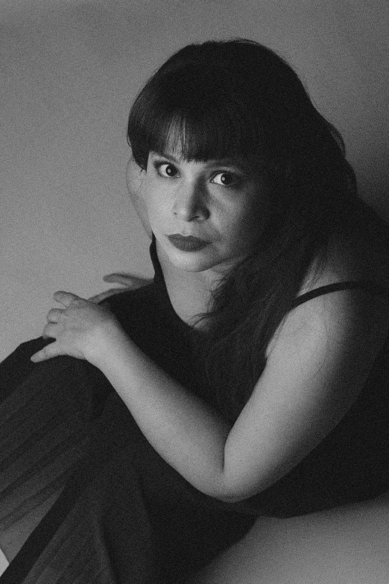 Retrato en blanco y negro de la artistas con vestido oscuro viendo a la cámara sobre ella mientras está sentada.